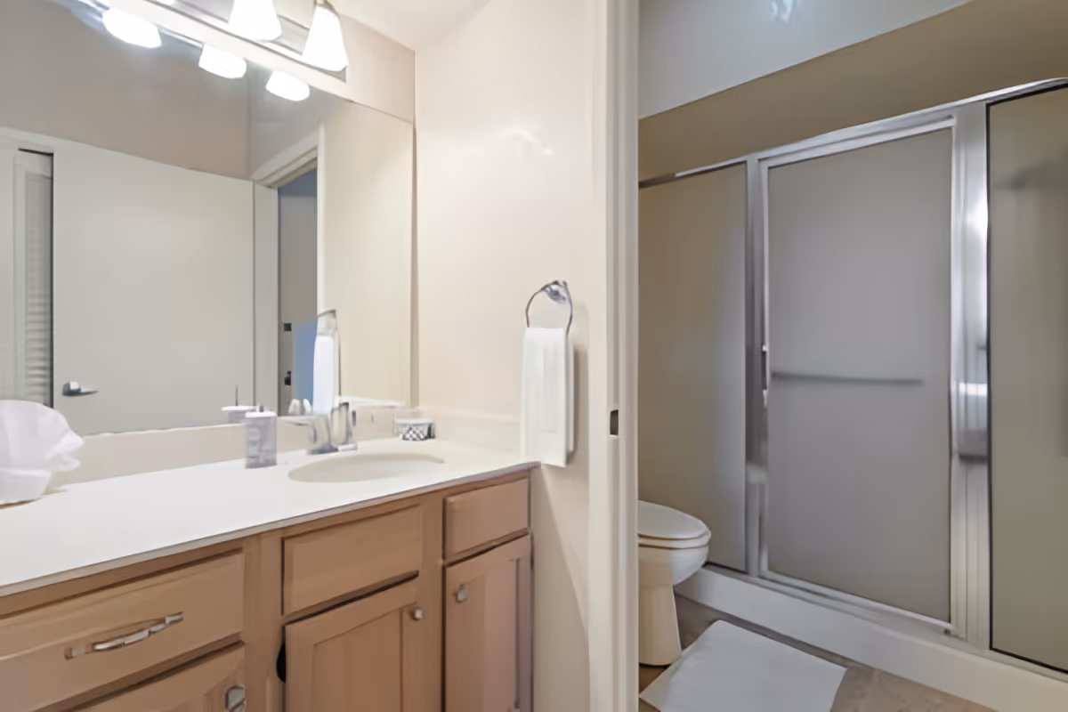 A clean bathroom with a wooden vanity featuring a white countertop and sink. Above the vanity is a large mirror with three light fixtures. To the right, there is a toilet and a shower with frosted glass sliding doors. A white towel hangs on a ring on the wall next to the vanity.