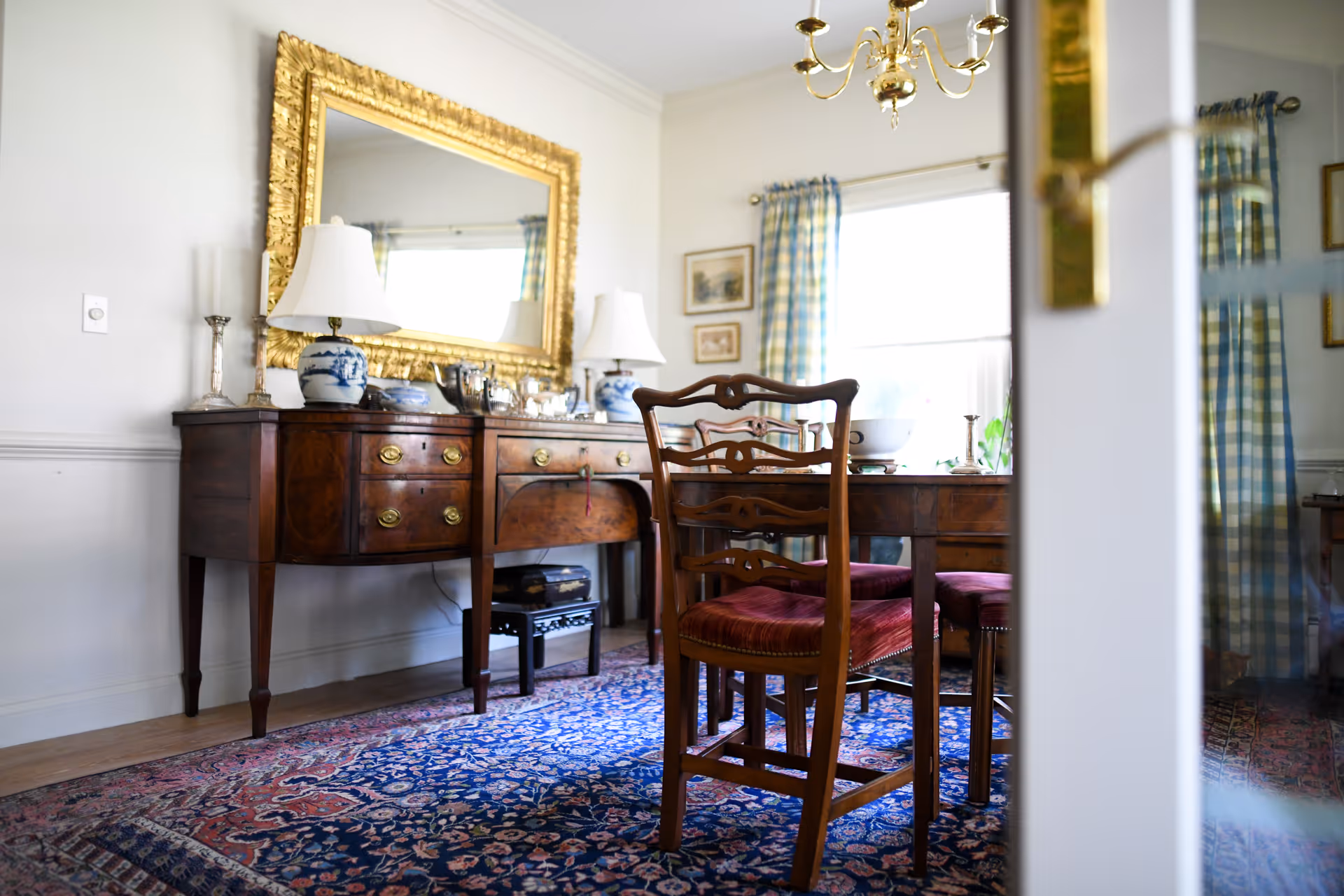 A traditional dining room with a wooden dining table and chairs with red cushions, a large ornate gold-framed mirror above a wooden sideboard, two white lamps with blue and white bases, and a patterned rug covering the floor. The room has light-colored walls, a window with blue and white checkered curtains, and a brass chandelier hanging from the ceiling.