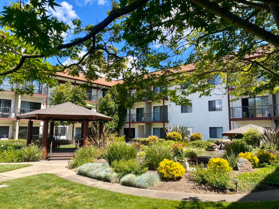 Outdoor garden area at Roseville Commons Senior Living with a gazebo, green grass, various shrubs and plants, and a three-story building with balconies in the background under a blue sky with some clouds.