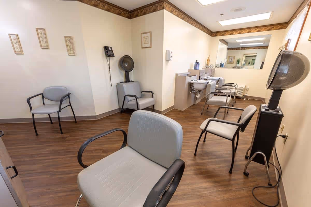 A small salon room with multiple styling chairs, hooded hair dryers, wash basins and a large mirror in a senior living facility.