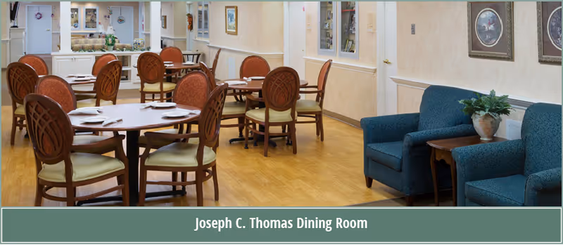 Dining room with round wooden tables and upholstered chairs and a small seating area with blue armchairs.