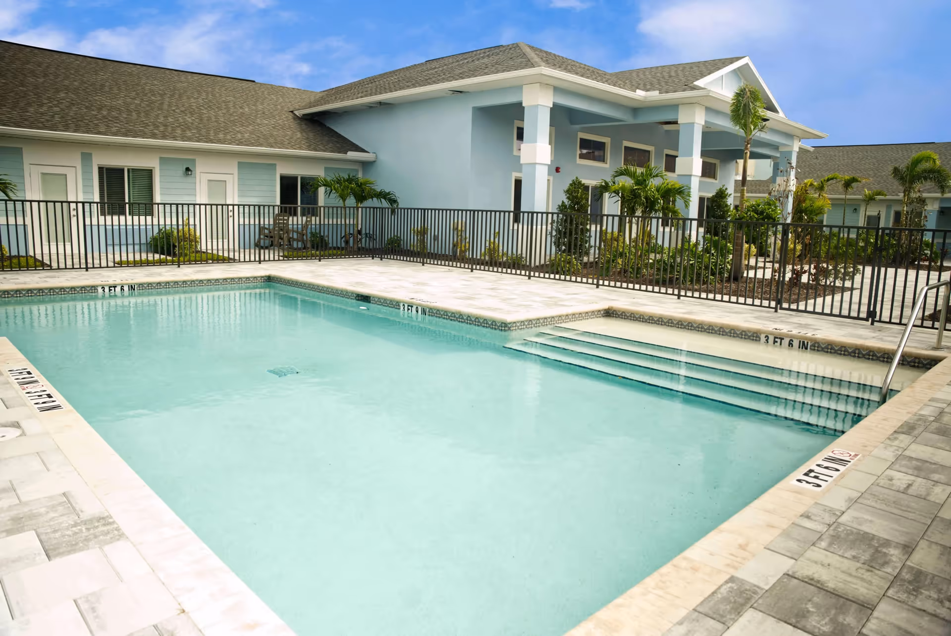 A clean outdoor swimming pool with steps and a safety fence in front of a light blue single-story assisted living building with palm trees.