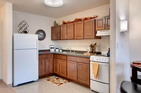 A compact kitchen with wooden cabinets, a white refrigerator, a white stove with a yellow towel hanging on the handle, a countertop with a sink, and decorative items including a clock, a wine rack, and autumn-themed garland on top of the cabinets.