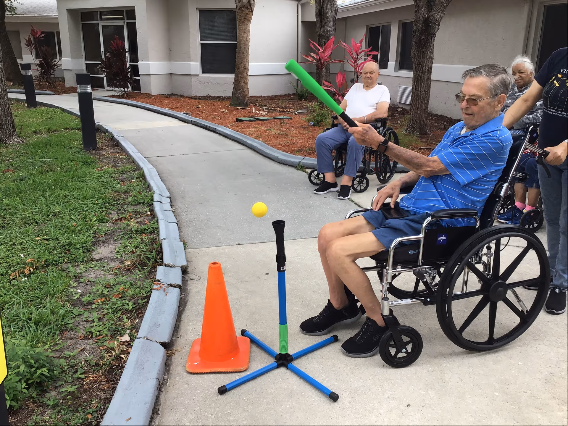 An elderly man in a wheelchair swings a green foam bat to hit a ball from a tee while other residents watch in a nursing home courtyard.