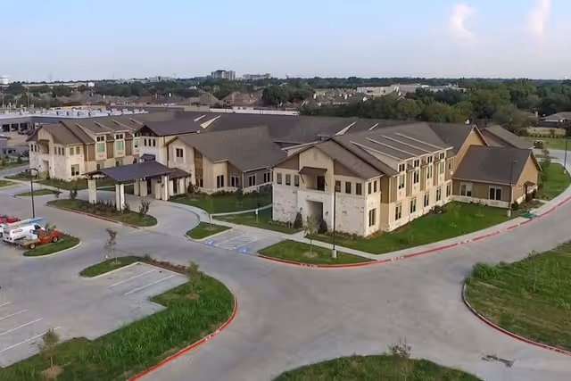 Aerial view of a large, modern senior living facility building with beige and stone exterior walls, multiple roof sections, and surrounding parking areas. The building is situated in a suburban area with greenery and other buildings in the background under a partly cloudy sky.