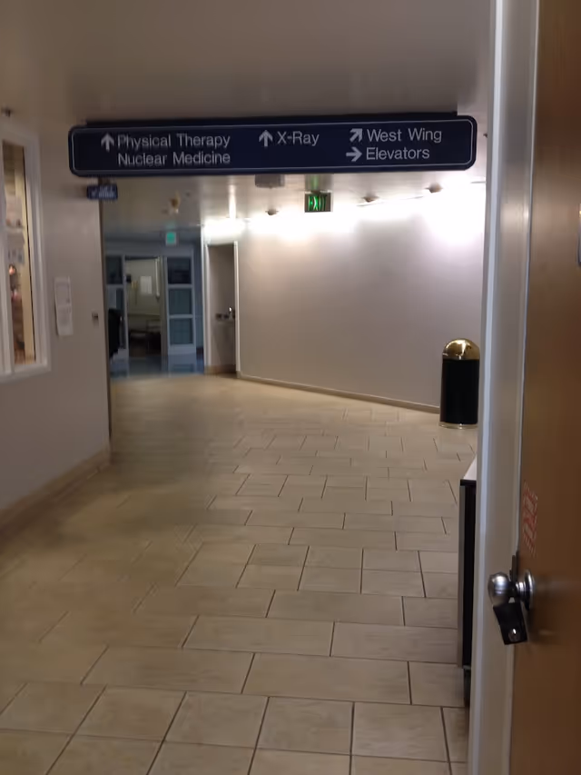 A hospital hallway with beige tiled floor and white walls. Overhead signs indicate directions to Physical Therapy, Nuclear Medicine, X-Ray, West Wing, and Elevators. There is a trash can with a gold-colored lid on the right side and a door partially visible on the right.