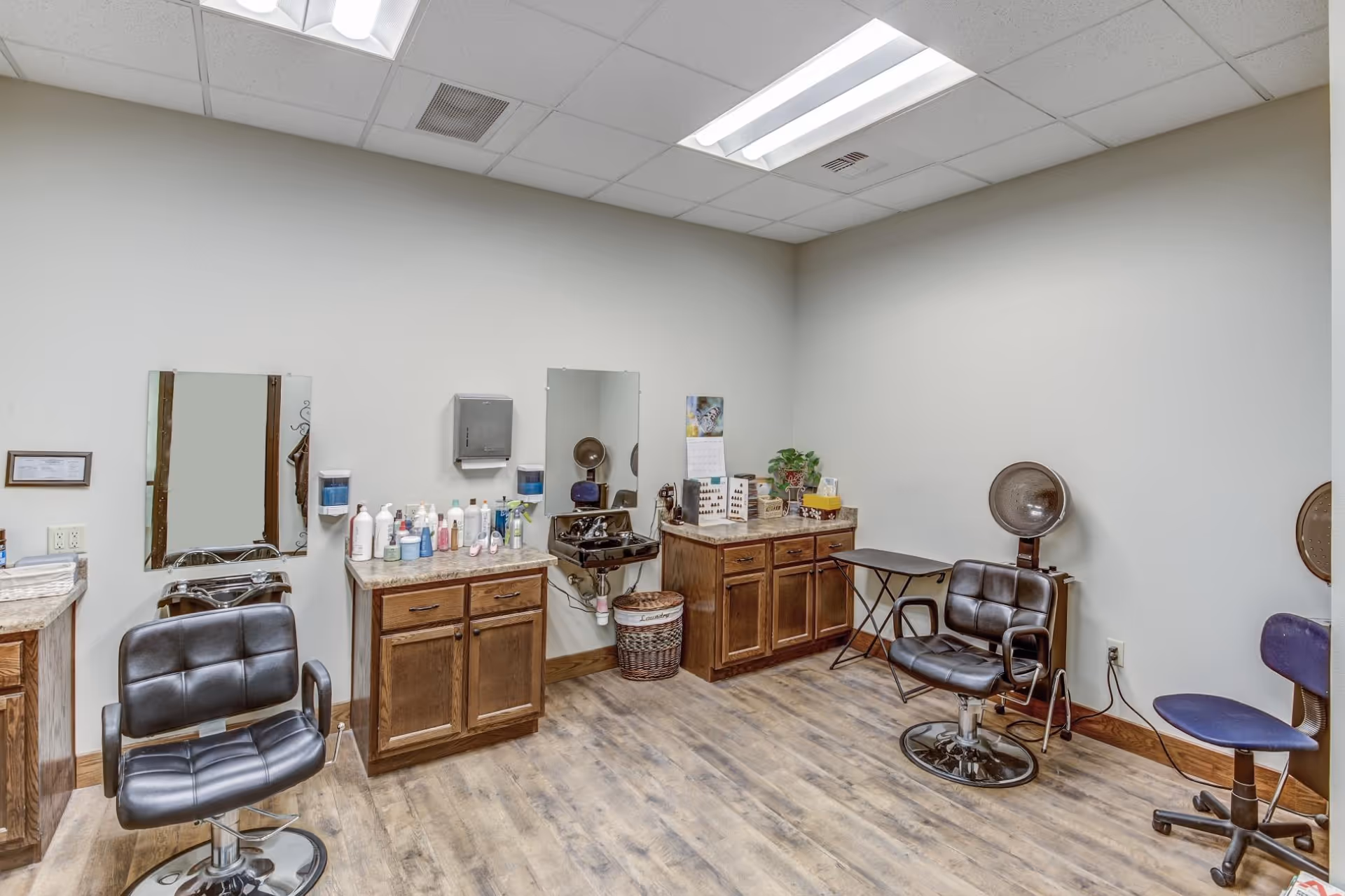 Small hair salon room with styling chairs, mirrors, cabinets stocked with hair products, and hooded dryers.