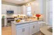 Bright and modern kitchen with white cabinets, a marble countertop island, stainless steel microwave and stove, and a small round dining table with chairs near a window with blinds. The kitchen is decorated with flowers and bowls of fruit.