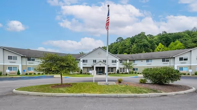 Front exterior view of a two-story senior living facility with a central entrance, an American flag on a flagpole in front, surrounded by landscaped greenery and a circular driveway.