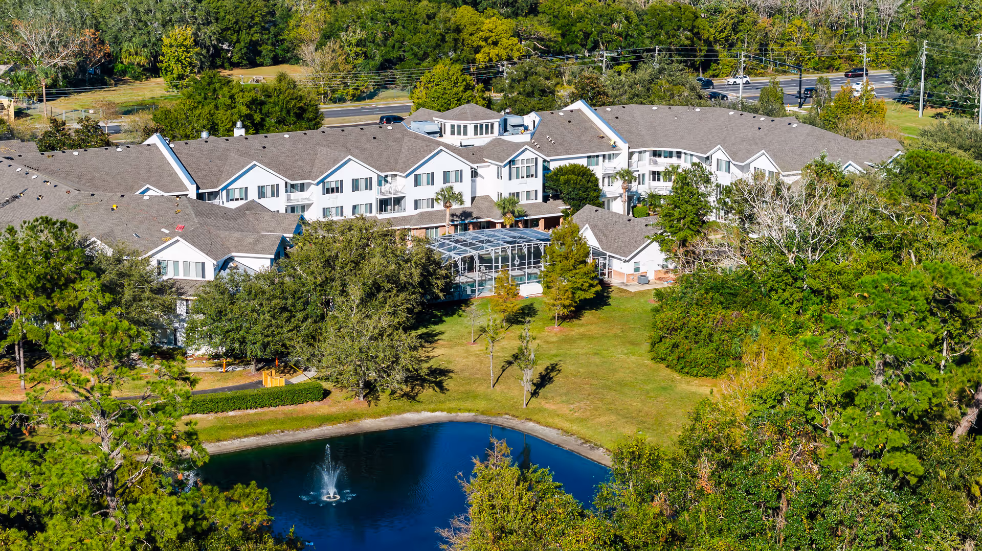 Aerial view of The Tremont - A Provincial Senior Living Community, showing a large multi-wing building surrounded by trees and greenery. In front of the building is a pond with a fountain, and a well-maintained lawn area. The building has white exterior walls and a gray roof, with a screened-in pool or patio area visible in the center.