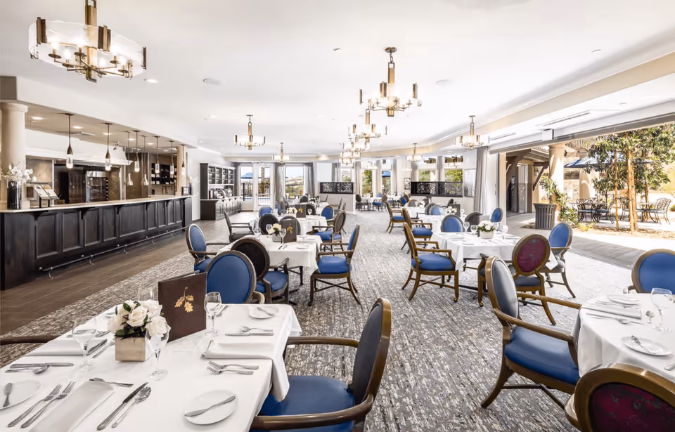 Spacious, elegant dining room with tables set in white linens and blue upholstered chairs, large windows and an outdoor patio visible.