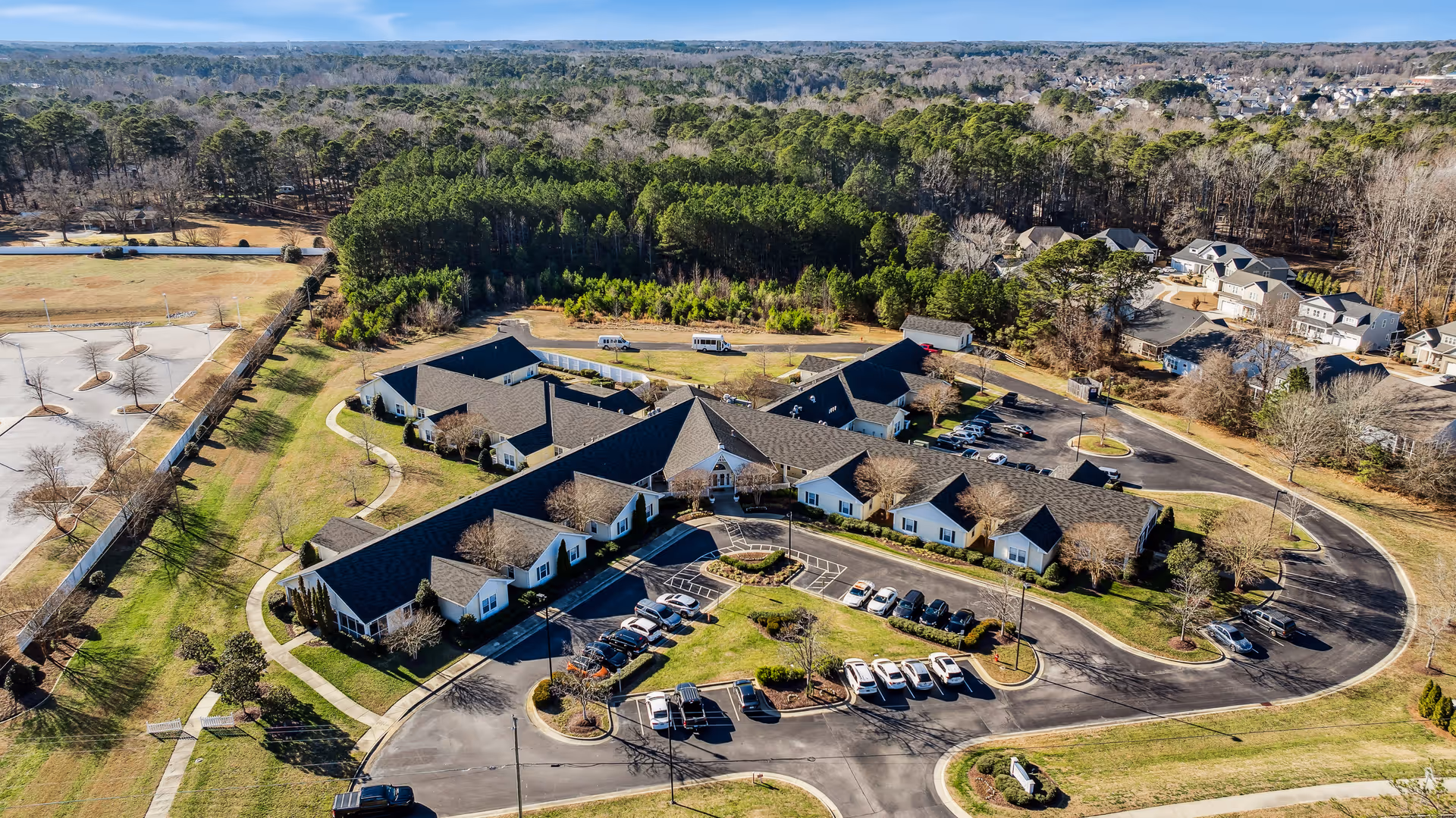 Aerial view of TerraBella Fuquay-Varina senior living facility showing a large single-story building with multiple wings surrounded by parking lots, landscaped areas, and trees. The facility is situated near a wooded area and residential neighborhood.