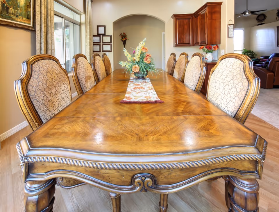 A long wooden dining table with ornate carved edges and eight upholstered chairs arranged around it. A floral centerpiece and a decorative table runner are placed in the middle of the table. The room has wooden flooring, a large window with curtains on the left, and wooden cabinets on the right. In the background, there is a living area with leather furniture and a ceiling fan.