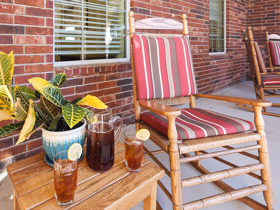 Wooden rocking chair with red, gray, and white striped cushions on a porch next to a small wooden table holding a potted plant and a pitcher with two glasses of iced tea garnished with lemon slices. The background shows a brick wall with windows.