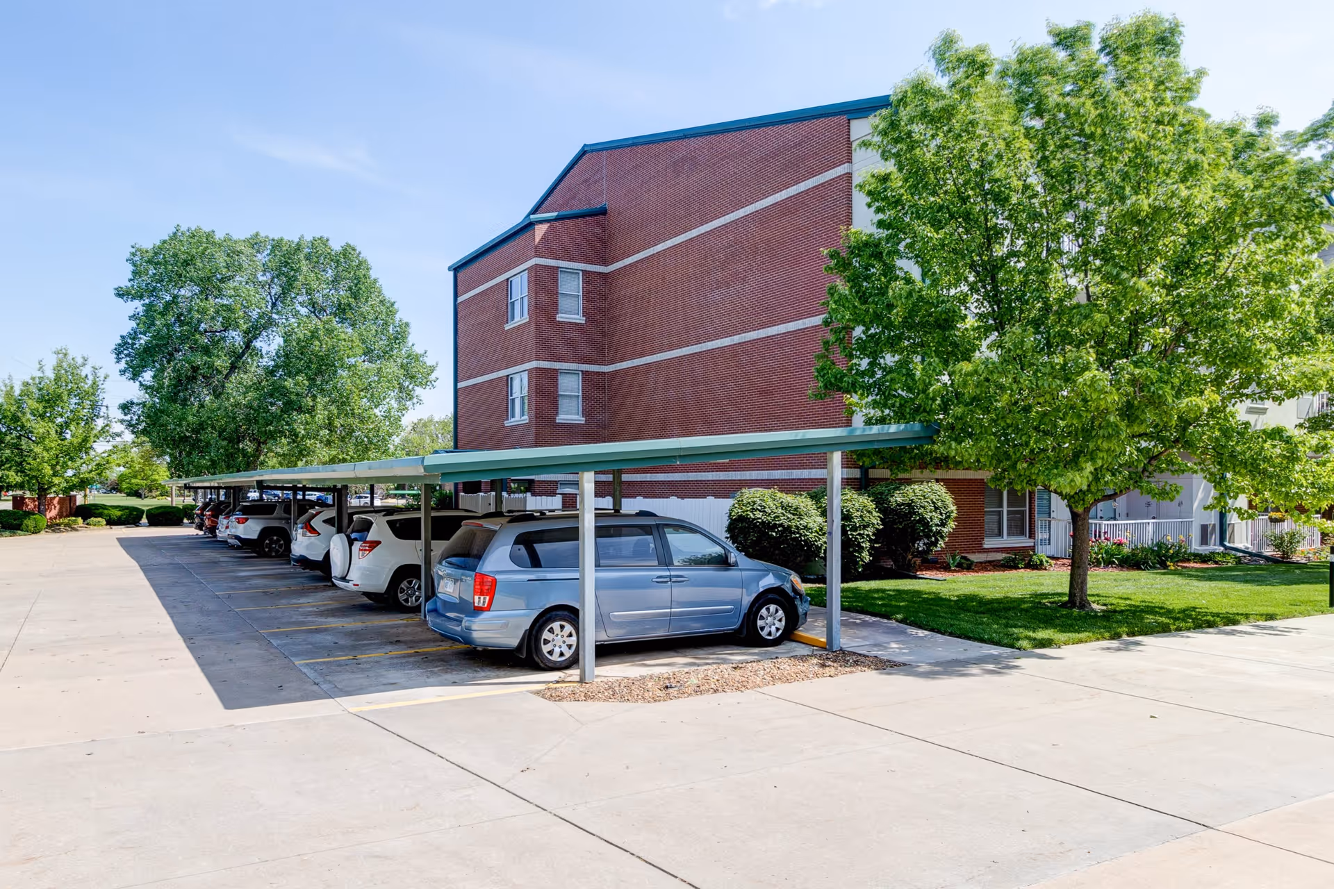 Parking area with a row of cars parked under a green metal carport next to a red brick building. There are green trees and bushes around the parking lot under a clear blue sky.