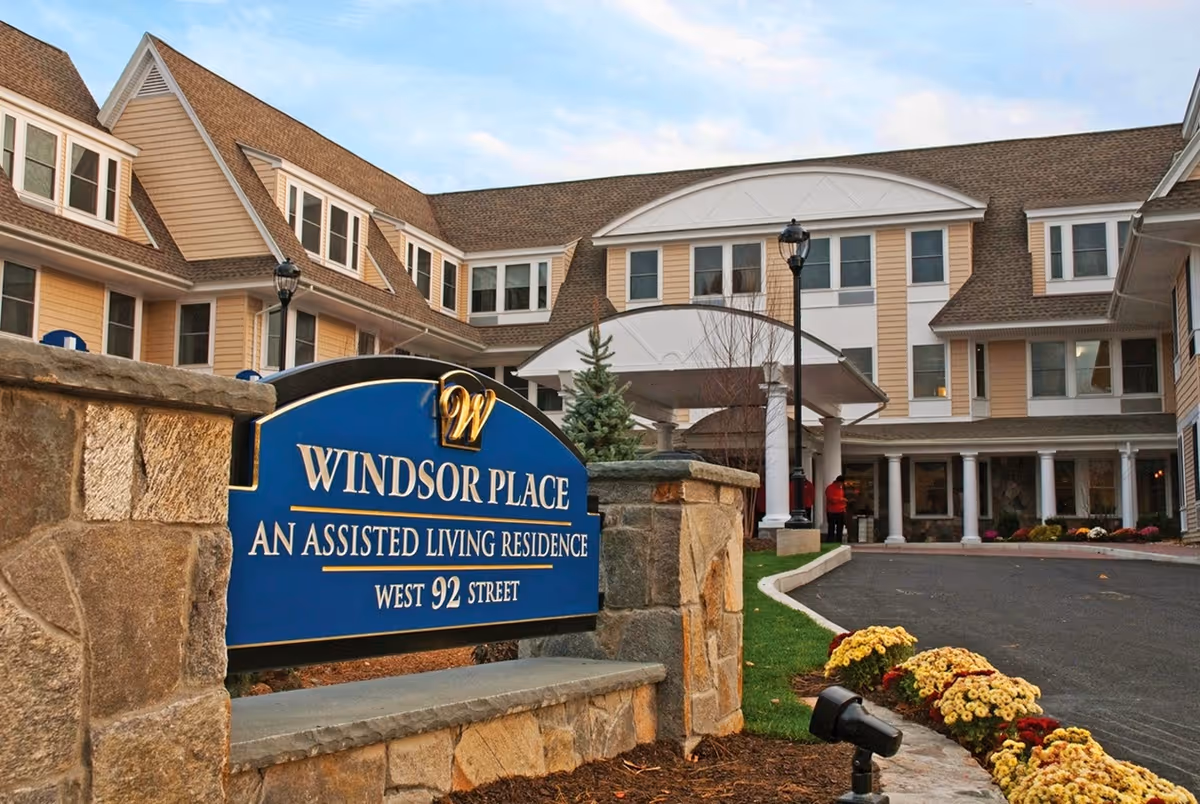 Exterior view of Windsor Place assisted living residence showing a large building with multiple windows and a peaked roof. In the foreground, there is a blue sign with gold lettering that reads 'Windsor Place An Assisted Living Residence West 92 Street,' surrounded by stone pillars and landscaping with flowers and a small tree.