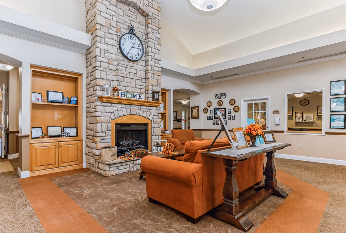 A cozy living room area in a senior living facility featuring a stone fireplace with a large clock above it. Two orange upholstered sofas face each other with a wooden table behind one sofa displaying framed pictures and a vase with orange flowers. The room has beige walls, carpeted floors, built-in wooden shelves with certificates, and decorative wall art including a sign that says 'Goodness Here'.