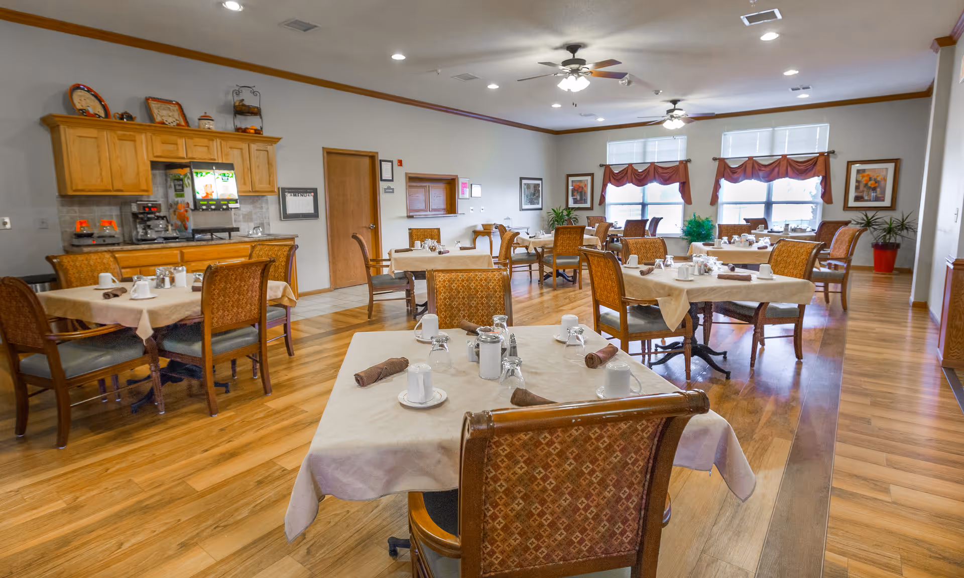 A dining room in a senior living facility with several tables covered in beige tablecloths, each set with cups, glasses, and rolled napkins. The room has wooden flooring, wooden chairs with patterned upholstery, and large windows with red valances letting in natural light. There is a wooden cabinet with coffee machines and a menu on the wall.