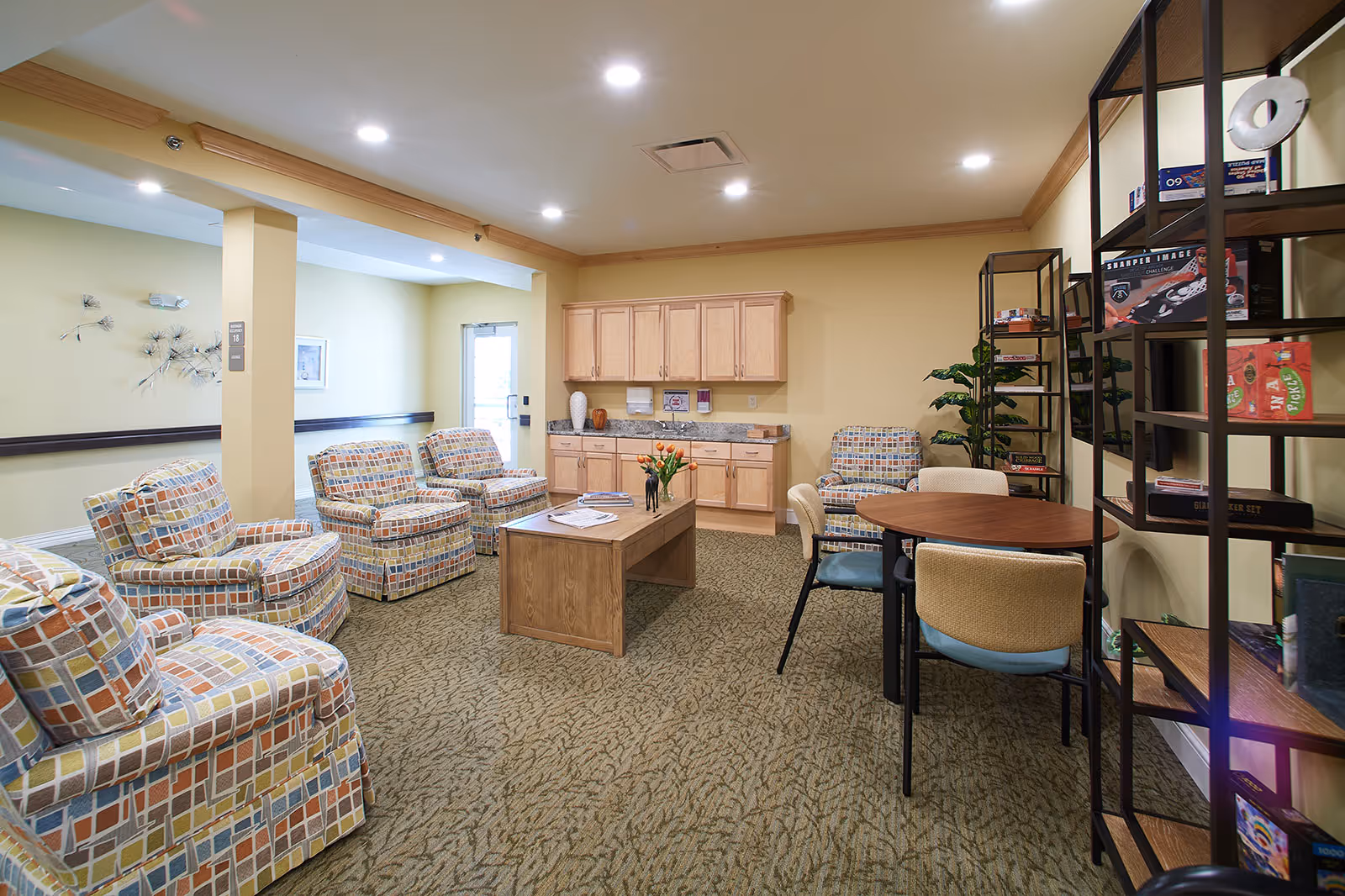 A cozy common area in a senior living facility with patterned armchairs arranged around a wooden coffee table, a small round table with chairs, a shelving unit with board games, and a cabinet with a countertop and decorative items against a beige wall.