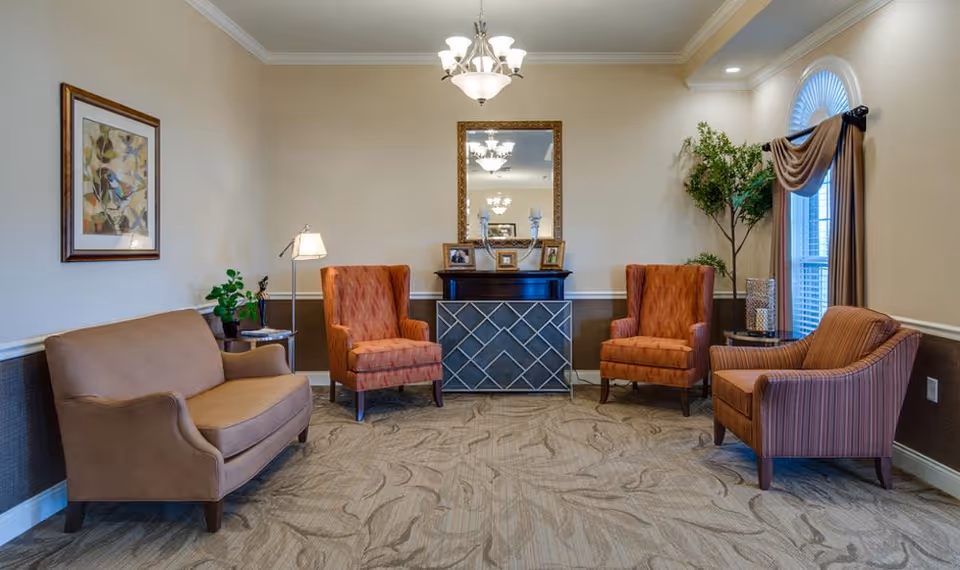 A cozy senior living facility lounge area with a beige loveseat, two orange armchairs, and a striped armchair arranged around a decorative console table with a large mirror above it. The room has beige walls, a patterned carpet, a window with drapes, a floor lamp, and some potted plants.