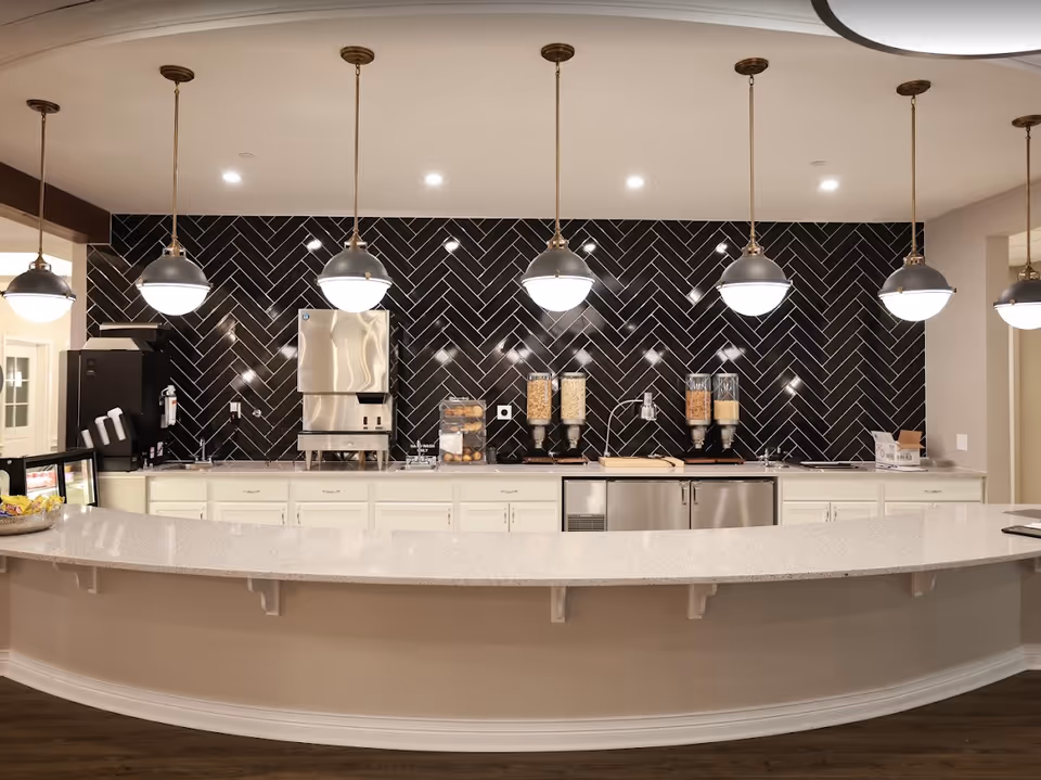 A modern kitchen area with a long curved white countertop and seven hanging pendant lights. The back wall features a black herringbone tile backsplash with stainless steel appliances, cereal dispensers, a coffee machine, and a display case with bagels. The floor is wooden and the cabinetry is white.