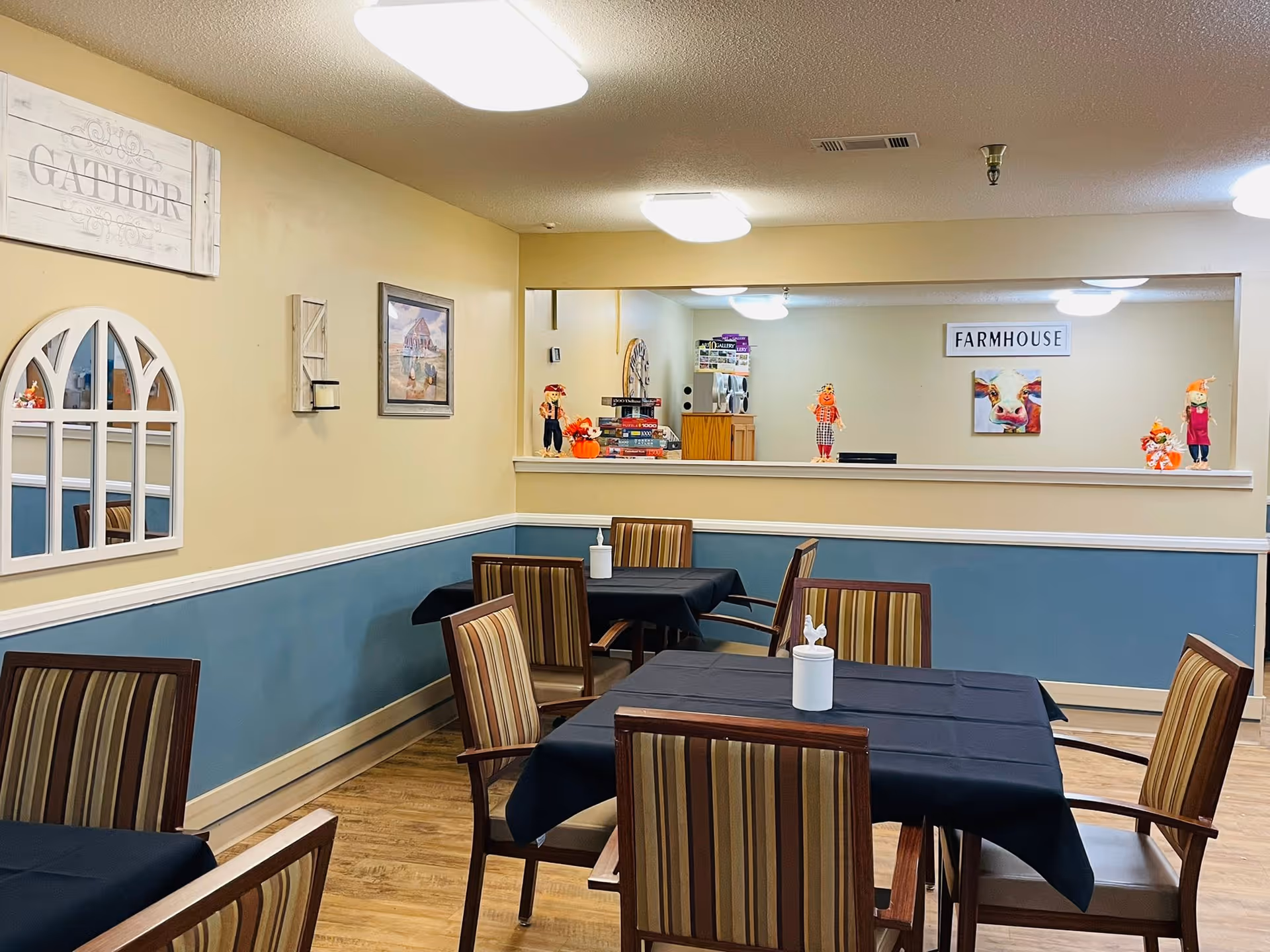 Communal dining room with several tables and striped chairs, farmhouse decor, and a serving pass-through window.