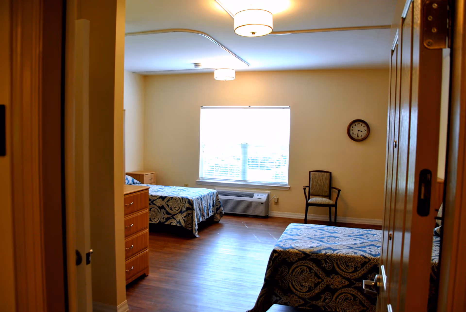 View into a bedroom with two twin beds covered in blue patterned bedspreads, a wooden dresser, a single chair against the wall, a window with blinds, and a wall clock showing 3:15. The room has wooden flooring and beige walls with ceiling lights.