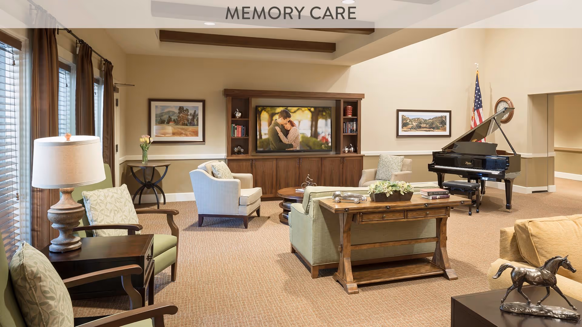 A spacious and well-lit memory care living room with comfortable seating including armchairs and sofas arranged around a wooden coffee table. A large flat-screen TV is mounted on a wooden entertainment center displaying a couple embracing. The room features a grand piano in the corner with an American flag behind it, framed artwork on the walls, and large windows with brown curtains allowing natural light to enter.