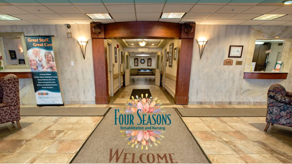 Lobby of the Four Seasons Rehabilitation and Nursing center with a logoed welcome mat, tiled floor, seating, reception desk, and a hallway leading inward.