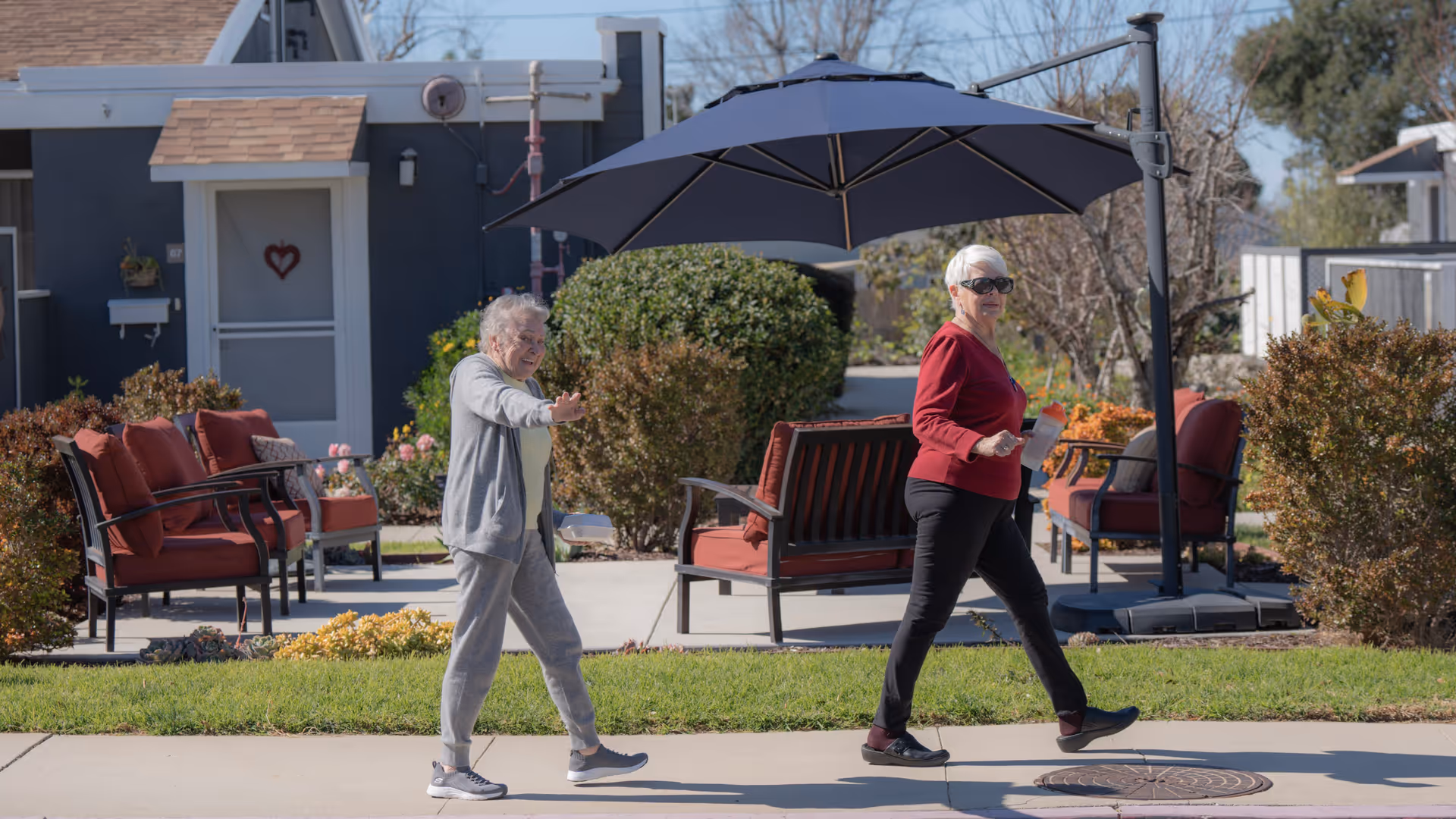 Two elderly women walking on a sidewalk in front of a residential building with outdoor seating and a large umbrella. One woman is wearing a gray outfit and waving, while the other is wearing a red top and black pants, holding a water bottle and wearing sunglasses. The area is landscaped with bushes and grass under a clear sky.