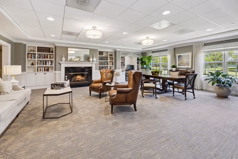 A spacious and well-lit living room with a beige carpet, two brown leather armchairs, a white sofa, a coffee table, and a fireplace with built-in bookshelves on either side. Large windows with white curtains allow natural light to fill the room, and there is a wooden dining table with chairs near the windows. The room is decorated with plants and framed artwork.