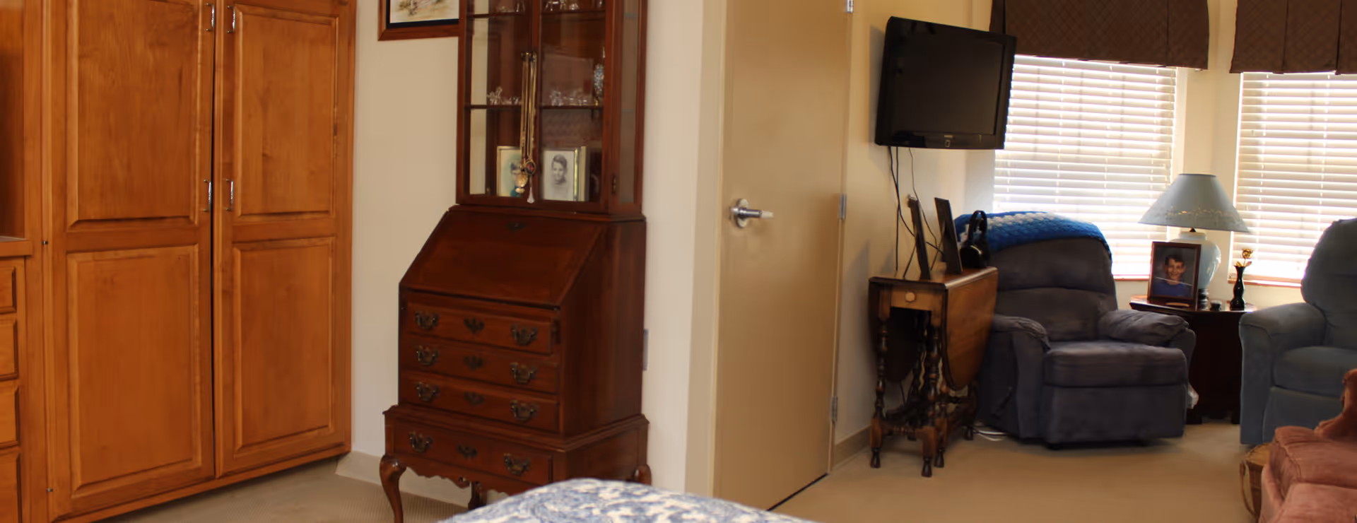 A cozy living room area with a wooden cabinet, a small antique desk with drawers, a wall-mounted TV, two comfortable armchairs, a side table with a lamp and framed photos, and windows with blinds and valances letting in natural light.