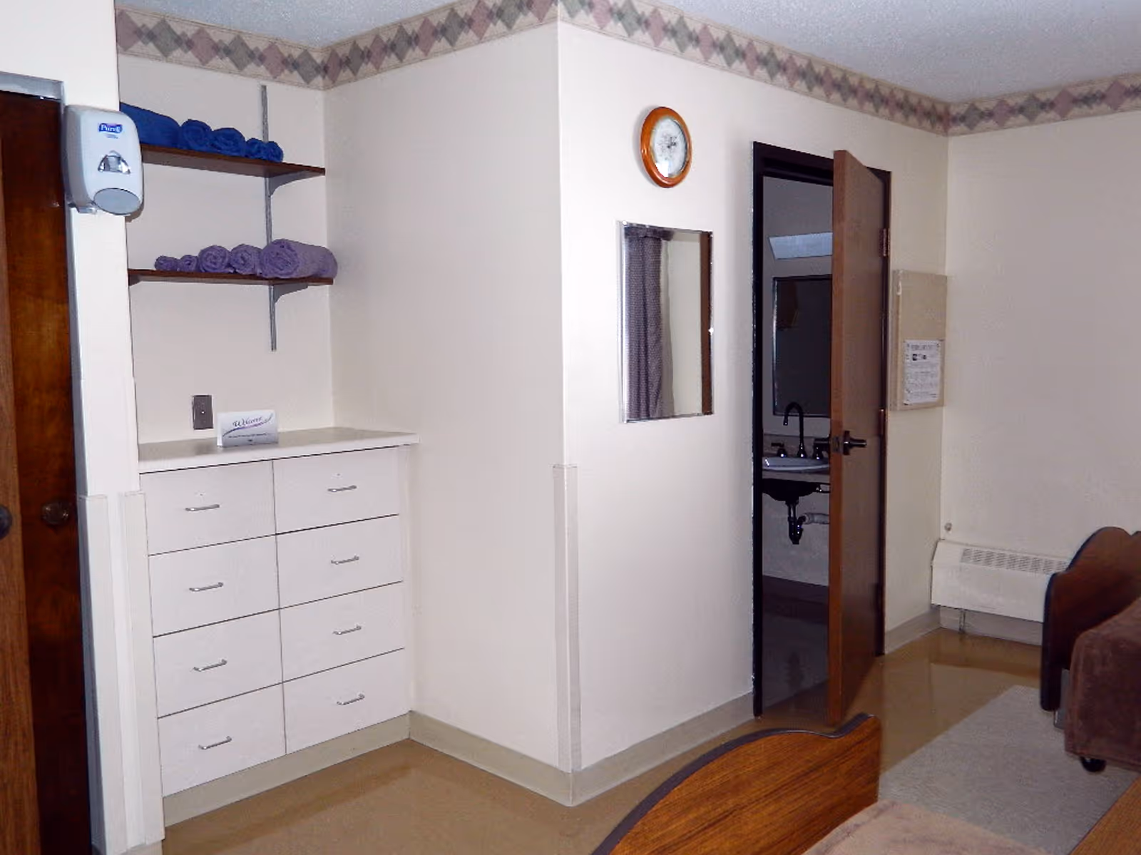 Interior view of a room in Bradford Manor showing a corner with white drawers and shelves holding folded towels. A wall-mounted soap dispenser is visible on the left. There is a clock and a mirror on the wall, and an open door reveals a sink in an adjacent bathroom. Part of a bed and a heater are also visible.