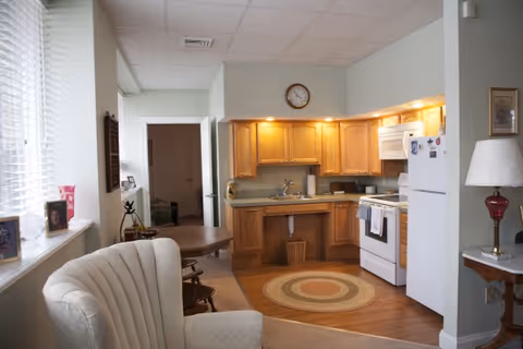 A cozy kitchen area with wooden cabinets, a white refrigerator, stove, and microwave. There is a round woven rug on the wooden floor in front of the sink. To the left, there is a small dining table with chairs and a comfortable upholstered armchair near a window with blinds. A clock is mounted on the wall above the cabinets, and a lamp with a red base is on a small table to the right.