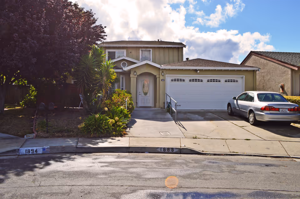 Front exterior view of a two-story residential building with a driveway, a white garage door, a silver car parked on the right side, and a front entrance with a white door. There are plants and a tree on the left side of the driveway, and the sky is partly cloudy.
