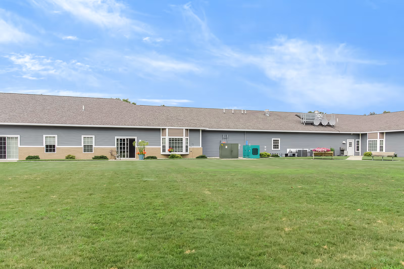 Exterior view of a single-story assisted living facility building with gray siding and a brown roof, surrounded by a large green lawn under a blue sky with some clouds. There are several windows, doors, and outdoor utility equipment visible along the building's wall.