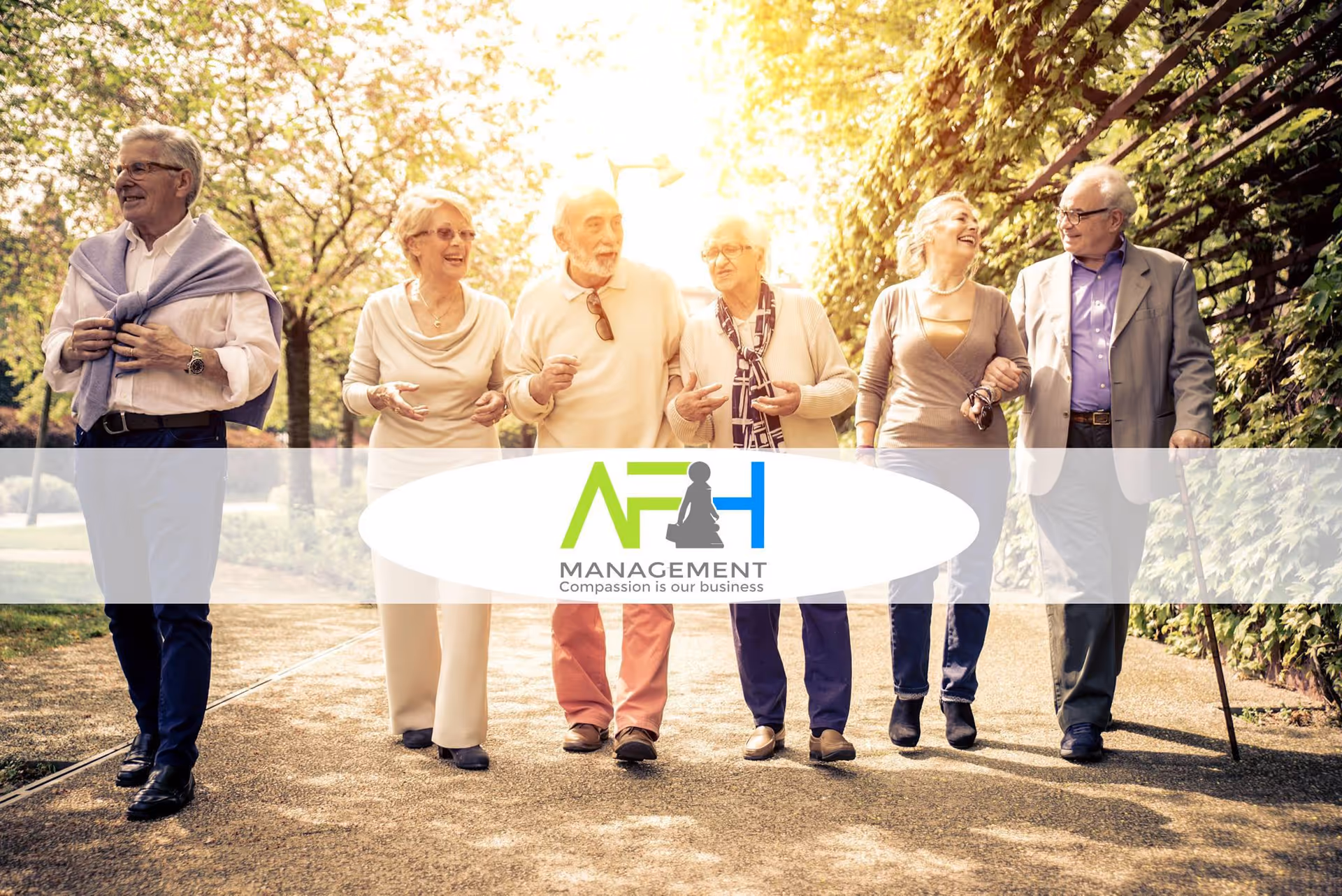 A group of six elderly people walking together outdoors on a sunny day, smiling and engaging with each other along a tree-lined path.