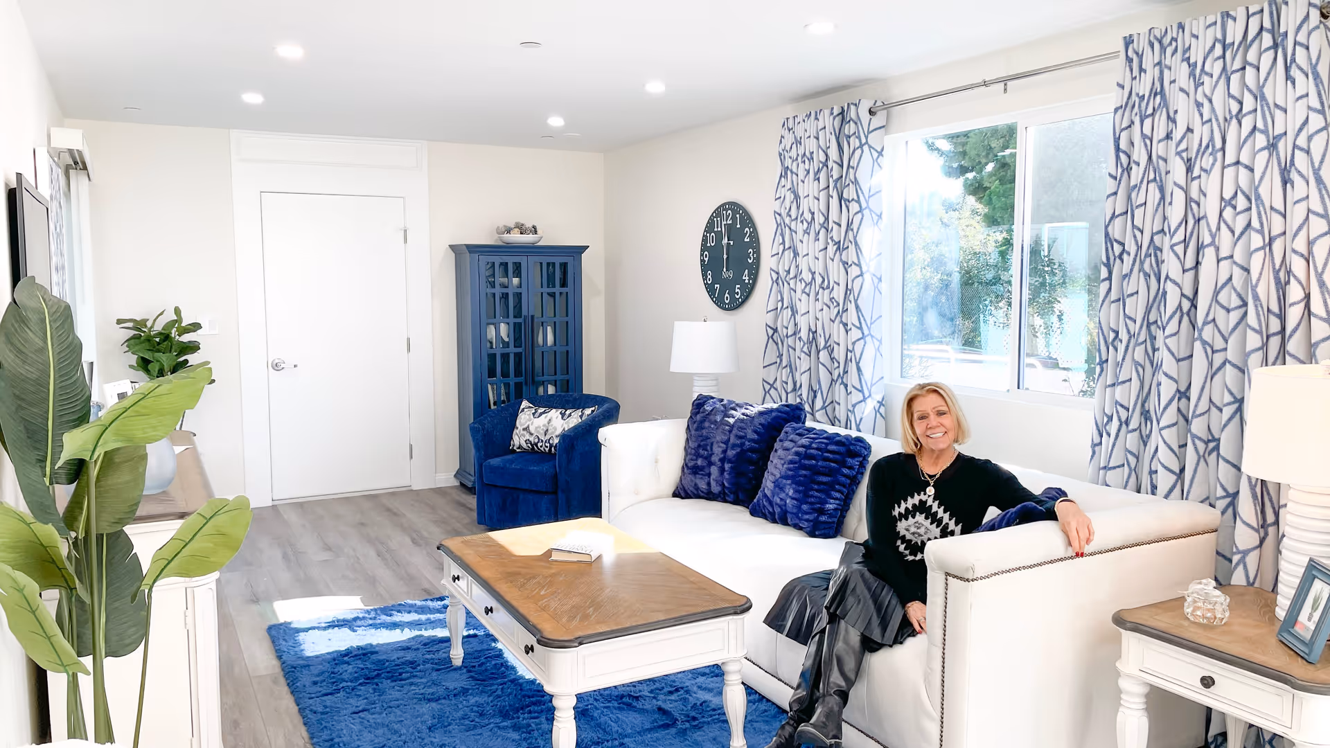 A bright living room with a white couch adorned with blue pillows, a wooden coffee table, a blue armchair, and a blue rug. A woman with blonde hair, wearing a black and white sweater and black boots, is sitting on the couch smiling. The room has light-colored walls, a large window with patterned blue and white curtains, a blue cabinet, a clock on the wall, and some plants.