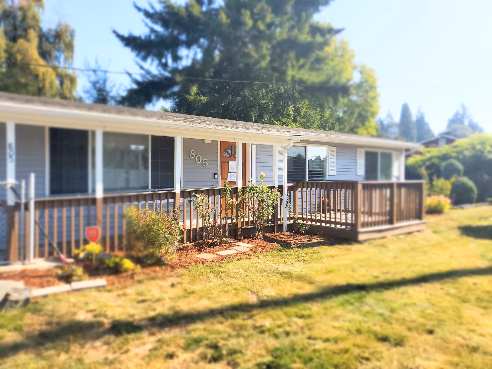 Single-story residential building with gray siding and white trim, featuring a wooden porch railing and a small garden with bushes and flowers in front. The house number 805 is displayed near the front door. Trees and greenery are visible in the background under a clear sky.
