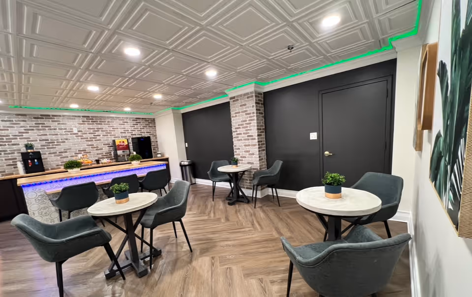 Communal dining/lounge area with round marble-top tables, upholstered chairs, a brick-accent service counter with beverage machines and LED lighting.