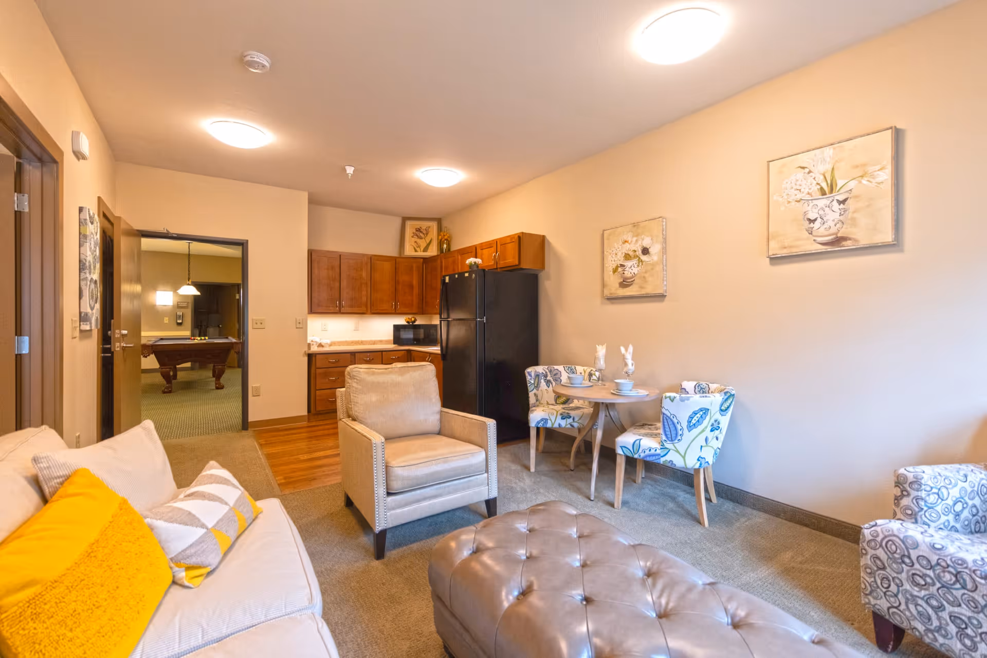 A cozy living area in a senior living facility featuring a beige sofa with yellow and patterned pillows, a beige armchair, a tufted leather ottoman, and a small round dining table with two floral upholstered chairs. The room has warm lighting, wooden cabinets, a black refrigerator, and two floral paintings on the wall. In the background, there is an open doorway leading to a room with a pool table.