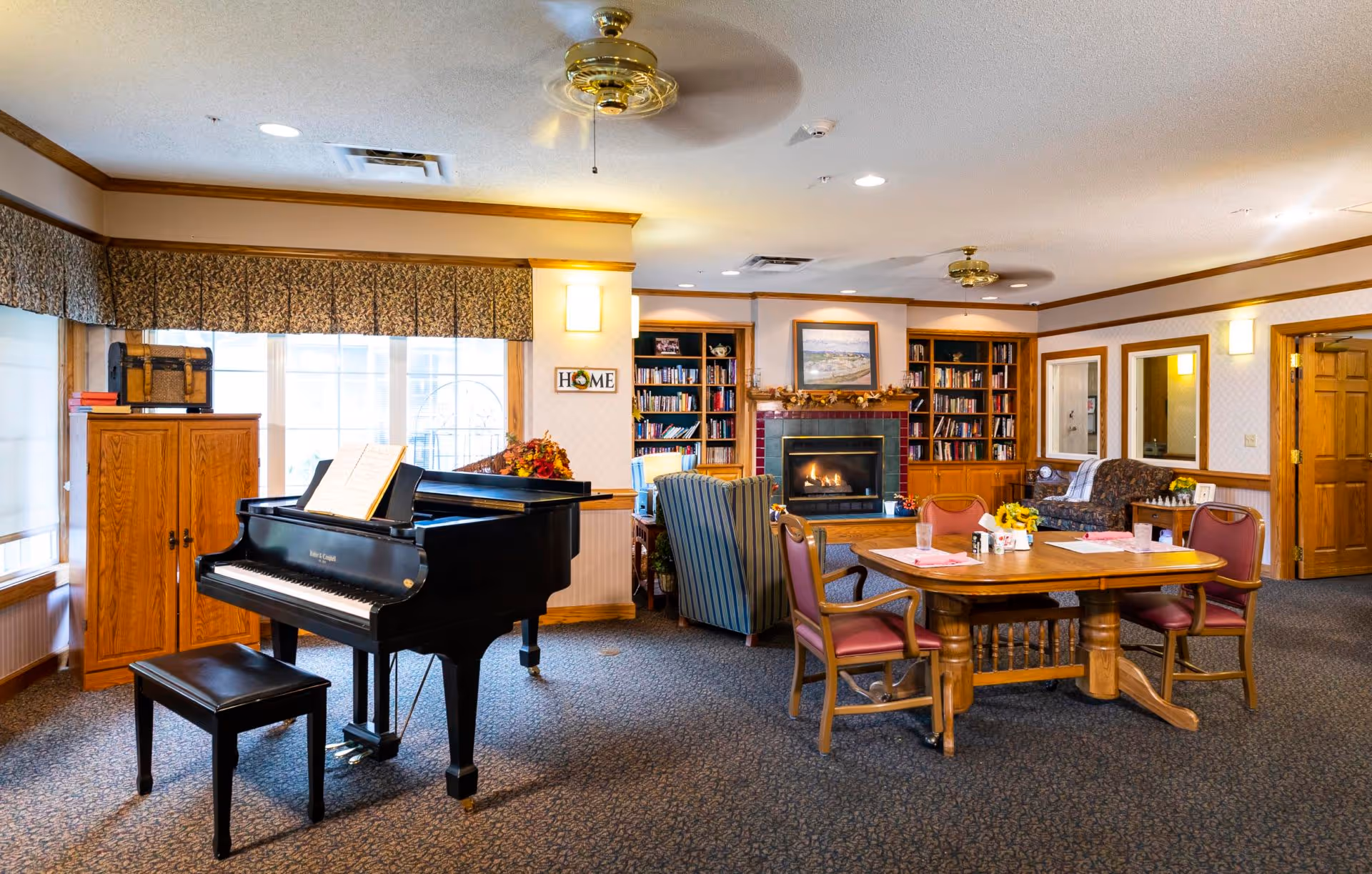 A cozy assisted-living common room with a grand piano, a table and chairs, and a seating area around a fireplace and bookshelves.