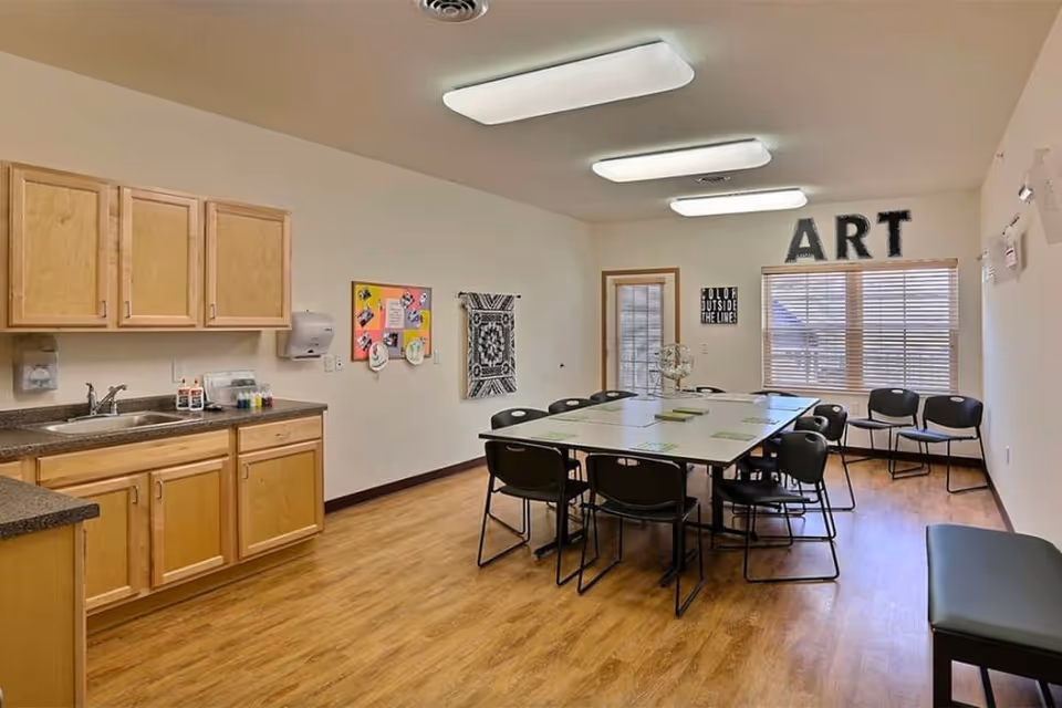 A well-lit art room with wooden flooring and light-colored walls. There is a large table in the center surrounded by black chairs. On the left side, there is a countertop with a sink and wooden cabinets above and below. The walls have various decorations including a colorful bulletin board, a woven wall hanging, and large letters spelling 'ART' above a window with blinds. There are also some chairs lined up against the wall and a door leading outside.