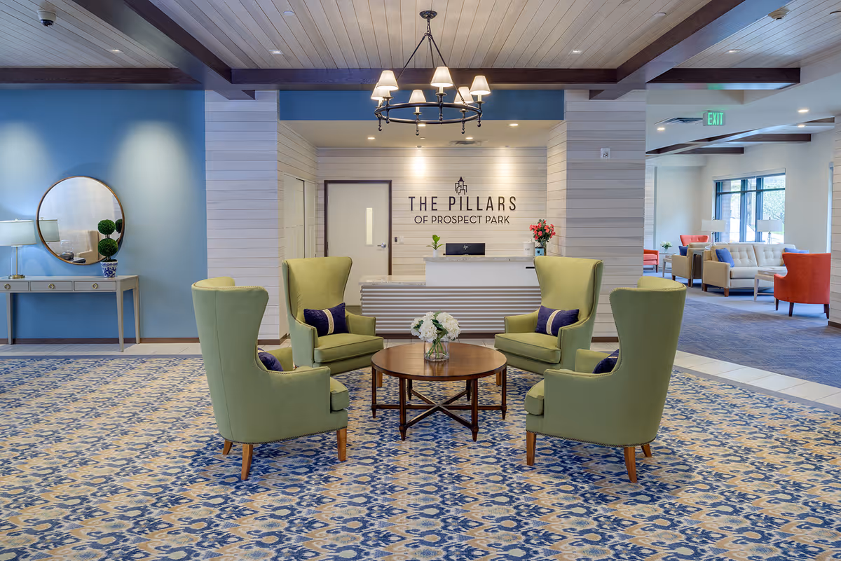 Bright reception lobby with four green wingback chairs around a round wooden table facing a white front desk with a sign reading 'The Pillars of Prospect Park'.
