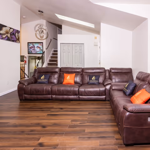 A spacious living room with dark brown leather sofas arranged in an L-shape, decorated with black and orange cushions bearing the Caring Arms logo. The room features wooden flooring, white walls, a skylight, a modern chandelier, and a staircase leading to an upper floor. A painting and a framed picture are visible on the walls.