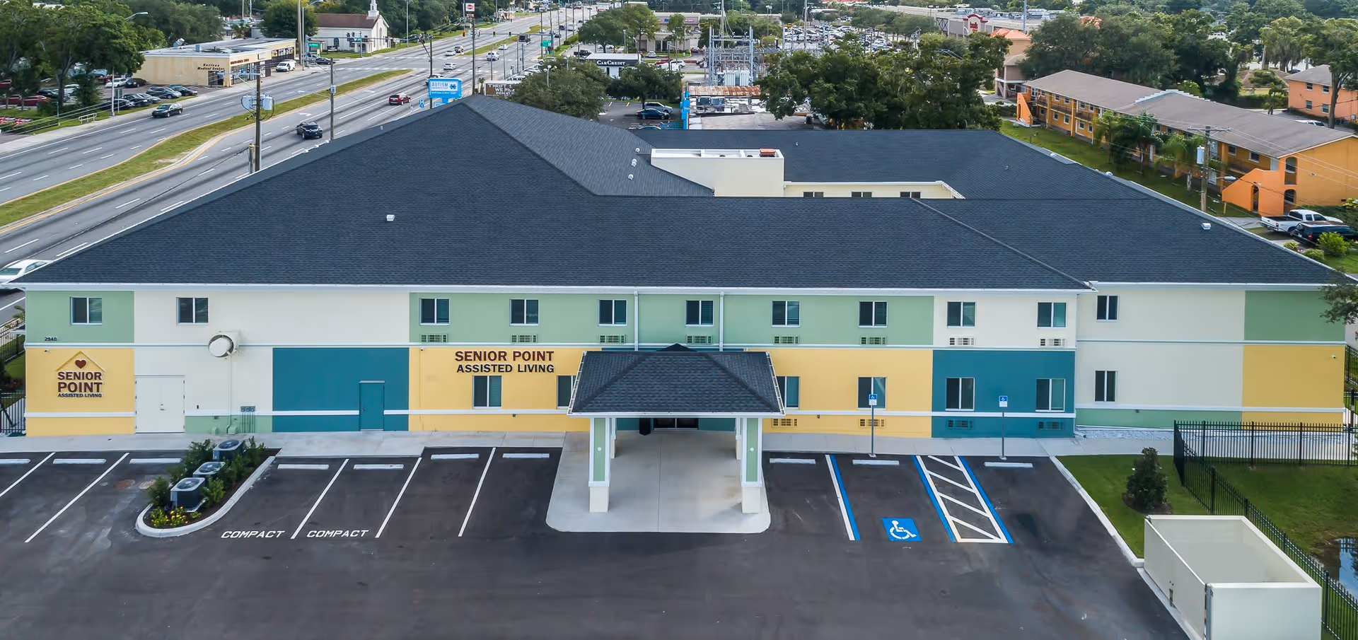 Front exterior view of Senior Point Assisted Living facility showing a two-story building with a covered entrance, multiple windows, and a parking lot with designated compact and handicapped parking spaces. The building is painted in white, green, and yellow colors and is located along a busy road with other buildings and trees in the background.