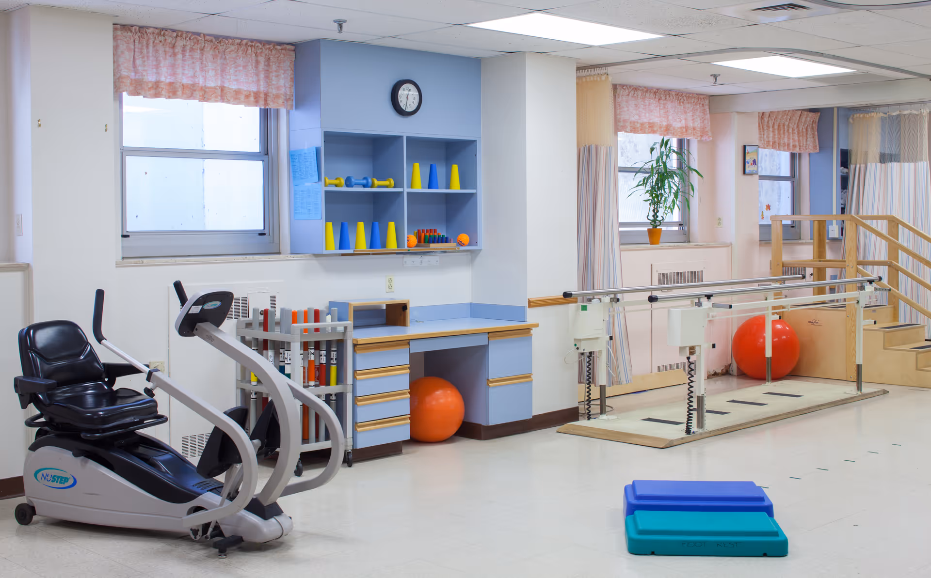 A physical therapy or rehabilitation room with exercise equipment including a recumbent bike, parallel bars for walking practice, colorful cones and weights on shelves, a small set of stairs with a handrail, and exercise steps on the floor. The room has windows with pink curtains and a potted plant on the windowsill.