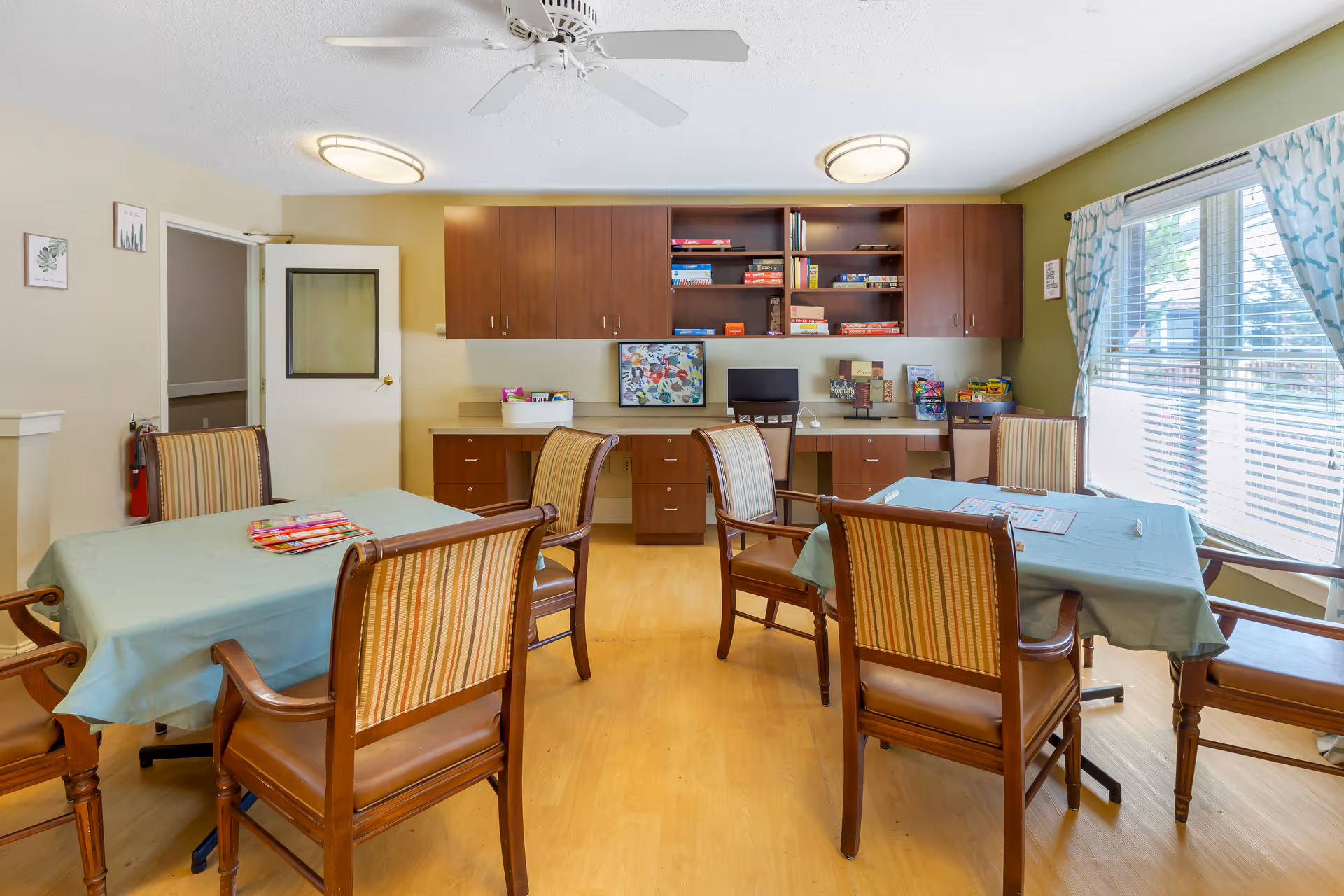A well-lit activity room with two tables covered in light blue tablecloths, each surrounded by wooden chairs with striped upholstery. The room has wooden flooring, a ceiling fan, and two round ceiling lights. On the back wall, there are wooden cabinets and shelves filled with board games and puzzles. A large window with blue patterned curtains lets in natural light.