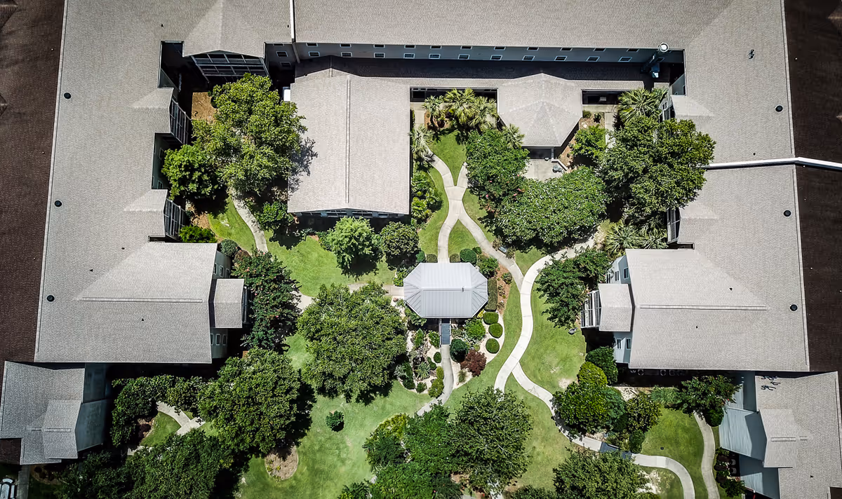 Aerial view of a senior living facility named Veranda of Pensacola showing a large building surrounding a landscaped garden with walking paths, trees, and a central gazebo.