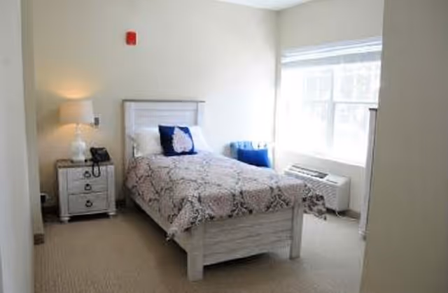 A small, bright bedroom with a single bed covered in a patterned bedspread and two pillows, one white and one blue. Next to the bed is a nightstand with a lamp and a telephone. A large window lets in natural light, and there is a blue chair and an air conditioning unit beneath the window. The walls are painted light beige and the floor is carpeted.